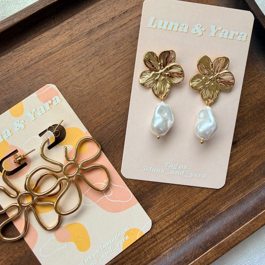 Gold floral earrings with pearl drops on a branded card, placed on a wooden surface.