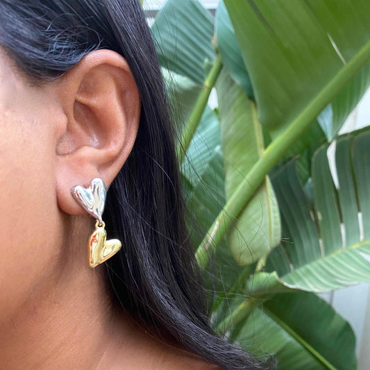 Close-up of an ear wearing a silver and gold heart-shaped earring with green leaves in the background.