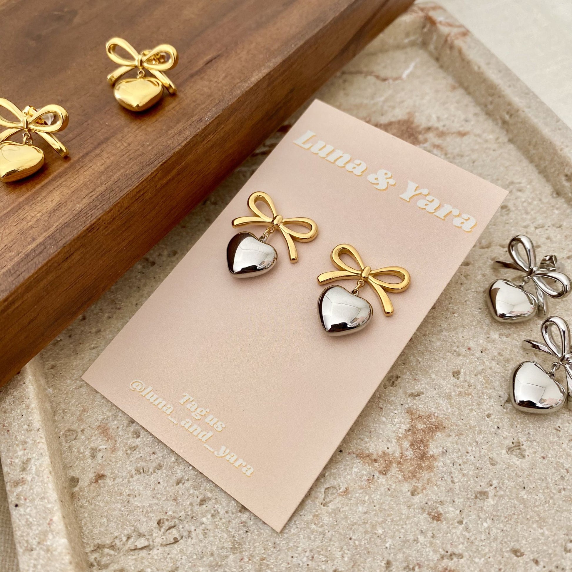 Gold and silver bow-shaped earrings on a Luna & Yara card with a wooden box in the background.